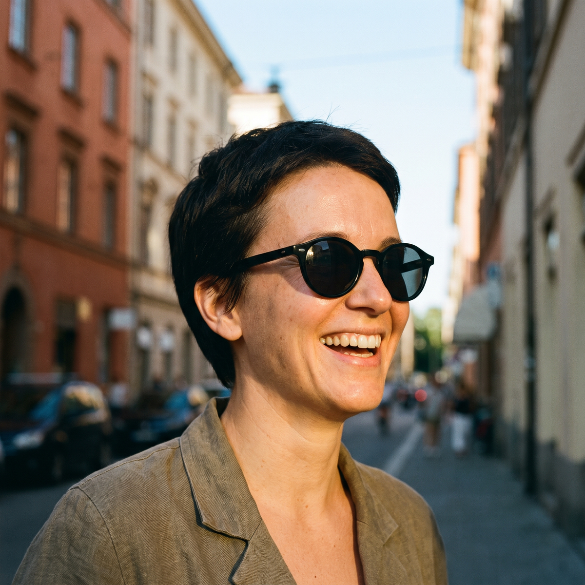 Woman with short dark hair wearing round black sunglasses on a sunlit street